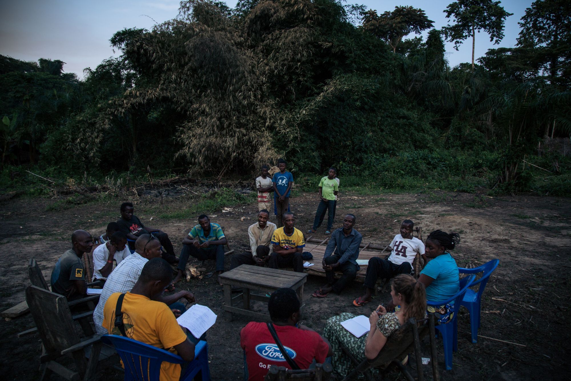 Some twenty people seated in a circle on bare earth at dusk, at the edge of the forest, taking notes during a village meeting.