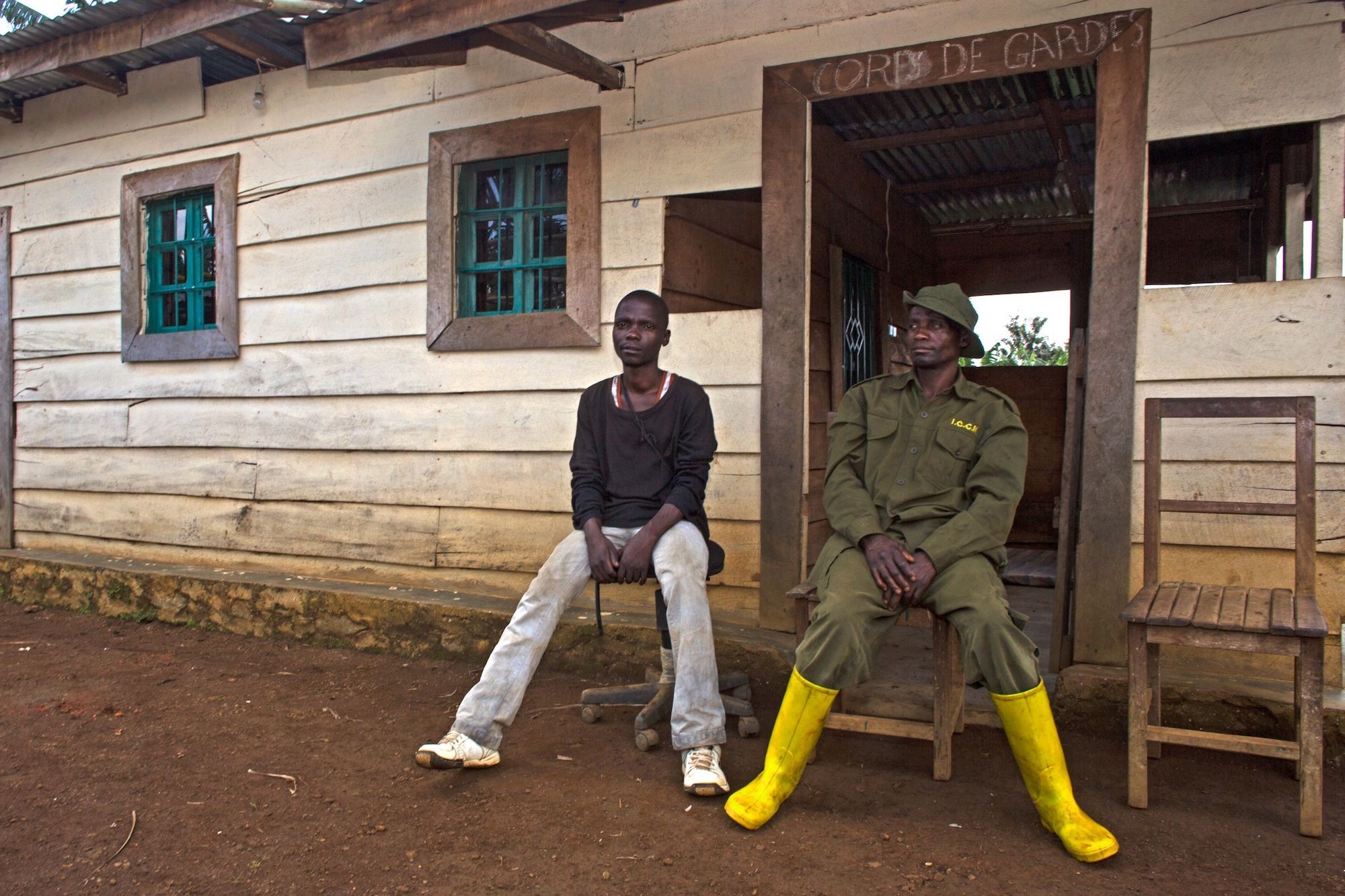 Two men seated in front of a wooden building marked “Corps de gardes”; one wears a ranger uniform and yellow rubber boots.