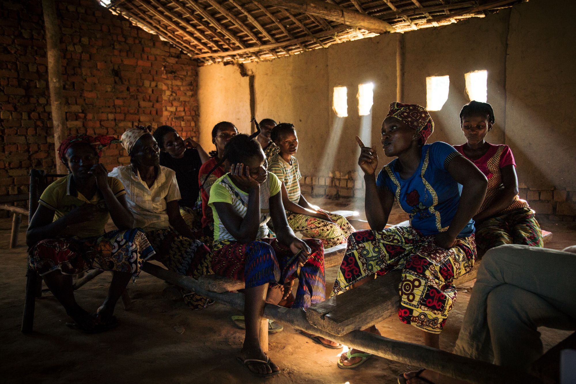 A group of women seated on wooden benches in a brick building lit by shafts of sunlight; one of them speaks and gestures.